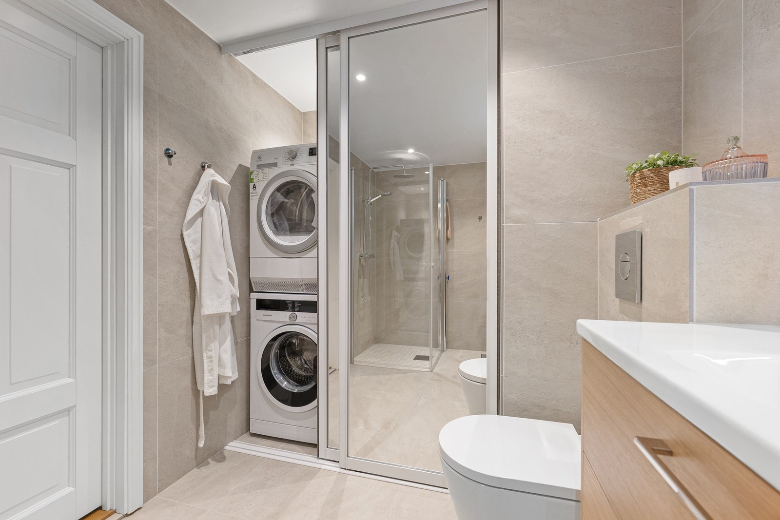 Praktisk og plassbesparende løsning med skyvedørsgarderobe som skjuler vaskemaskin og tørketrommel. Badet kombinerer komfort og funksjonalitet på en svært elegant måte. Galleribilde