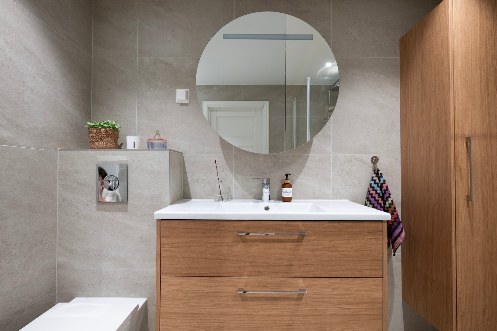 Lekker servantsone med heldekkende servant, moderne armatur og rundt speil med integrert belysning. Tidløse flater og varme detaljer i tre gir badet et eksklusivt og harmonisk uttrykk. Galleribilde