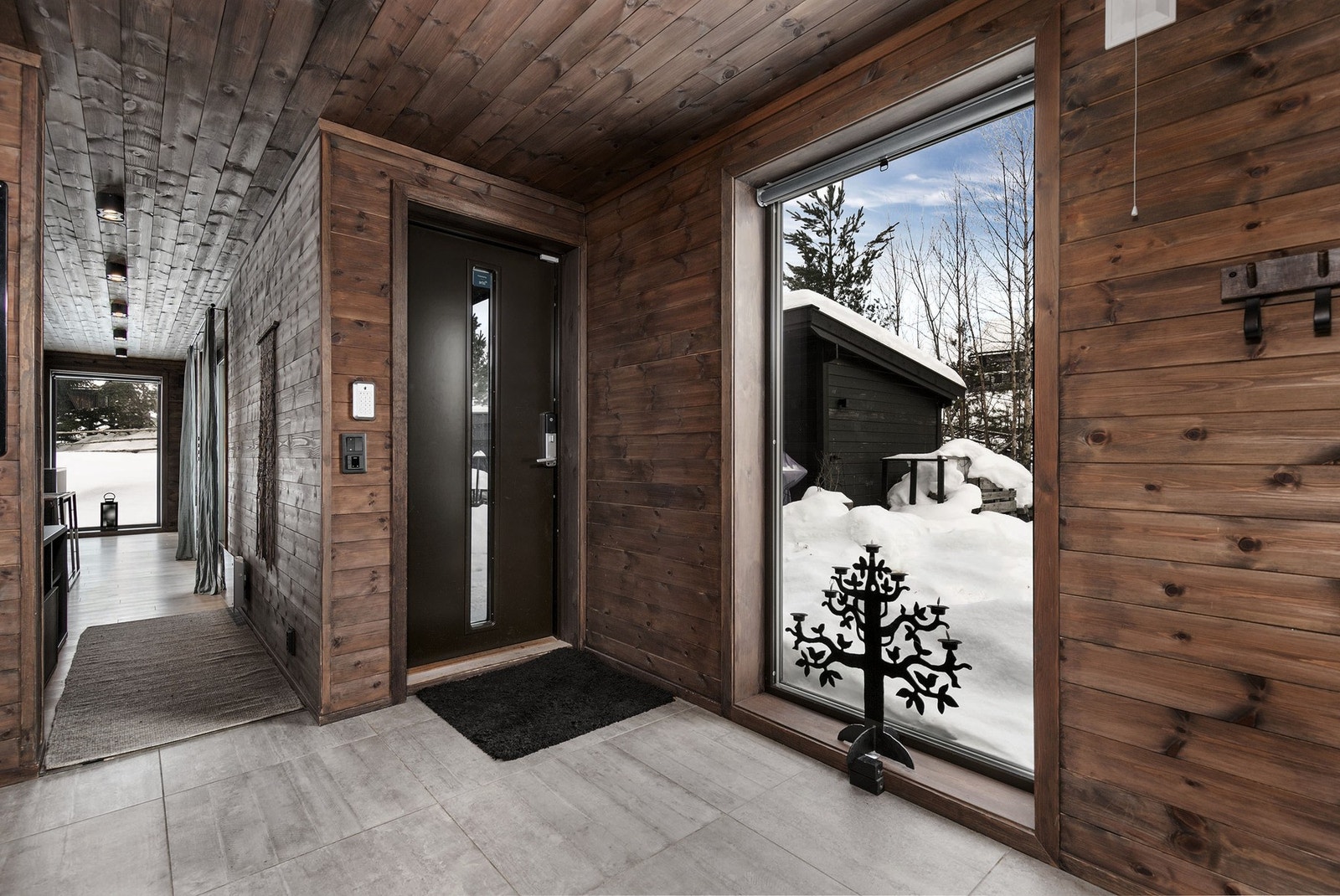 Romslig hall med storformatfliser på gulv med gulvvarme og et stort gulv-til-tak-vindu som gir et særdeles godt førsteinntrykk av fritidsboligen. Galleribilde