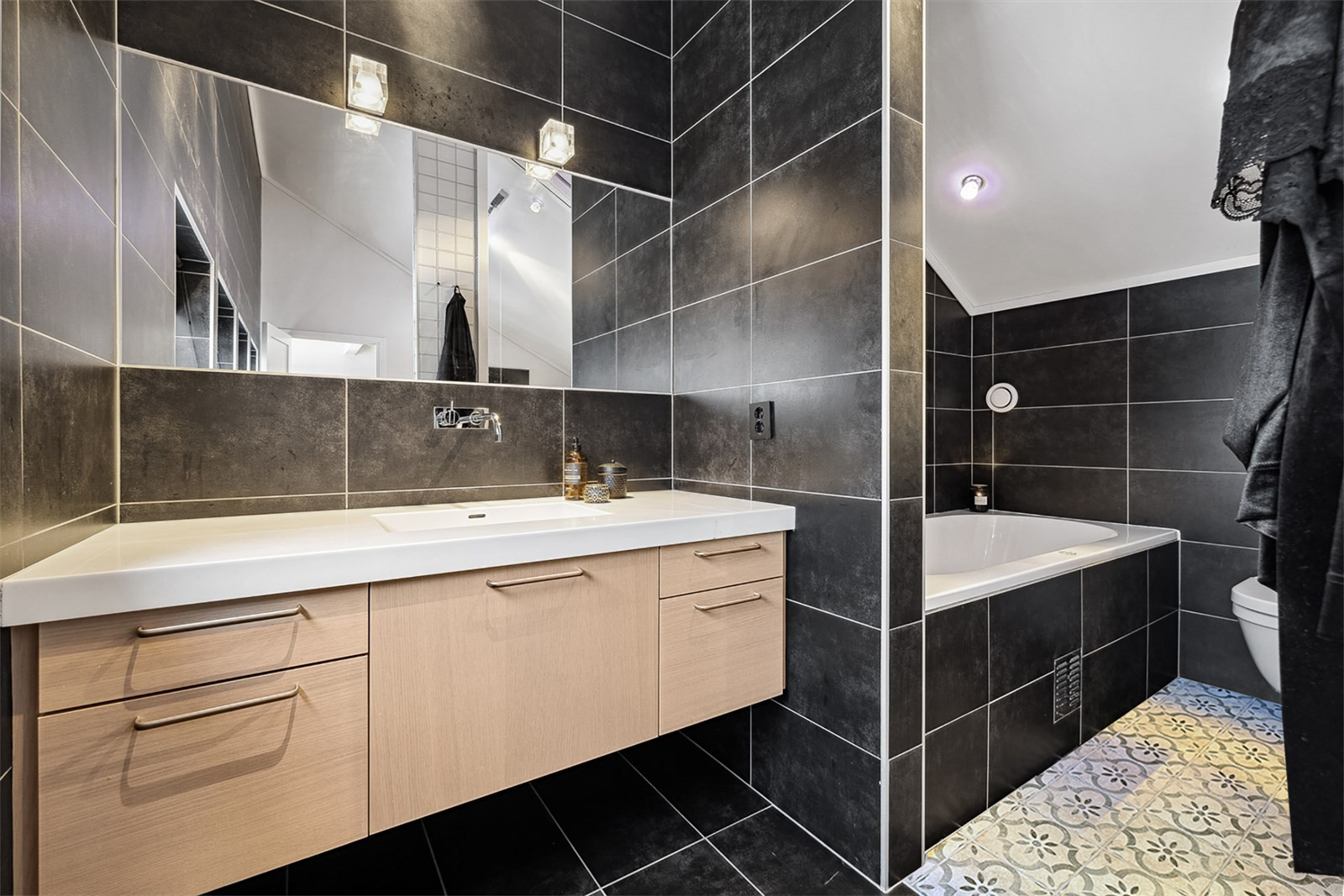 Flislagt bad med elektriske varmekabler i gulvet, innmurt badekar og vegghengt toalett. Galleribilde