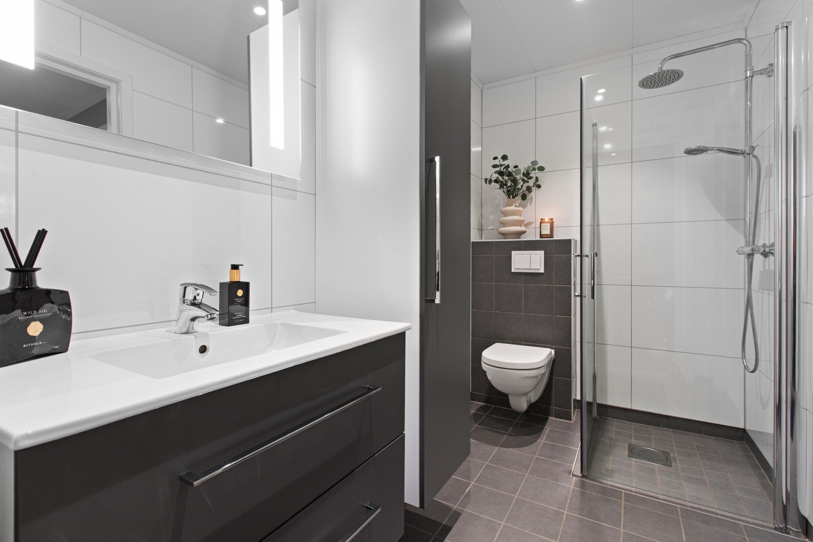 Badet er flislagt, har behagelig varme i gulvet og med himlingsplater i tak. Badet er innredet med vegghengt toalett, dusjhjørne med glassvegger, baderomsinnredning med servant og høyskap. Galleribilde