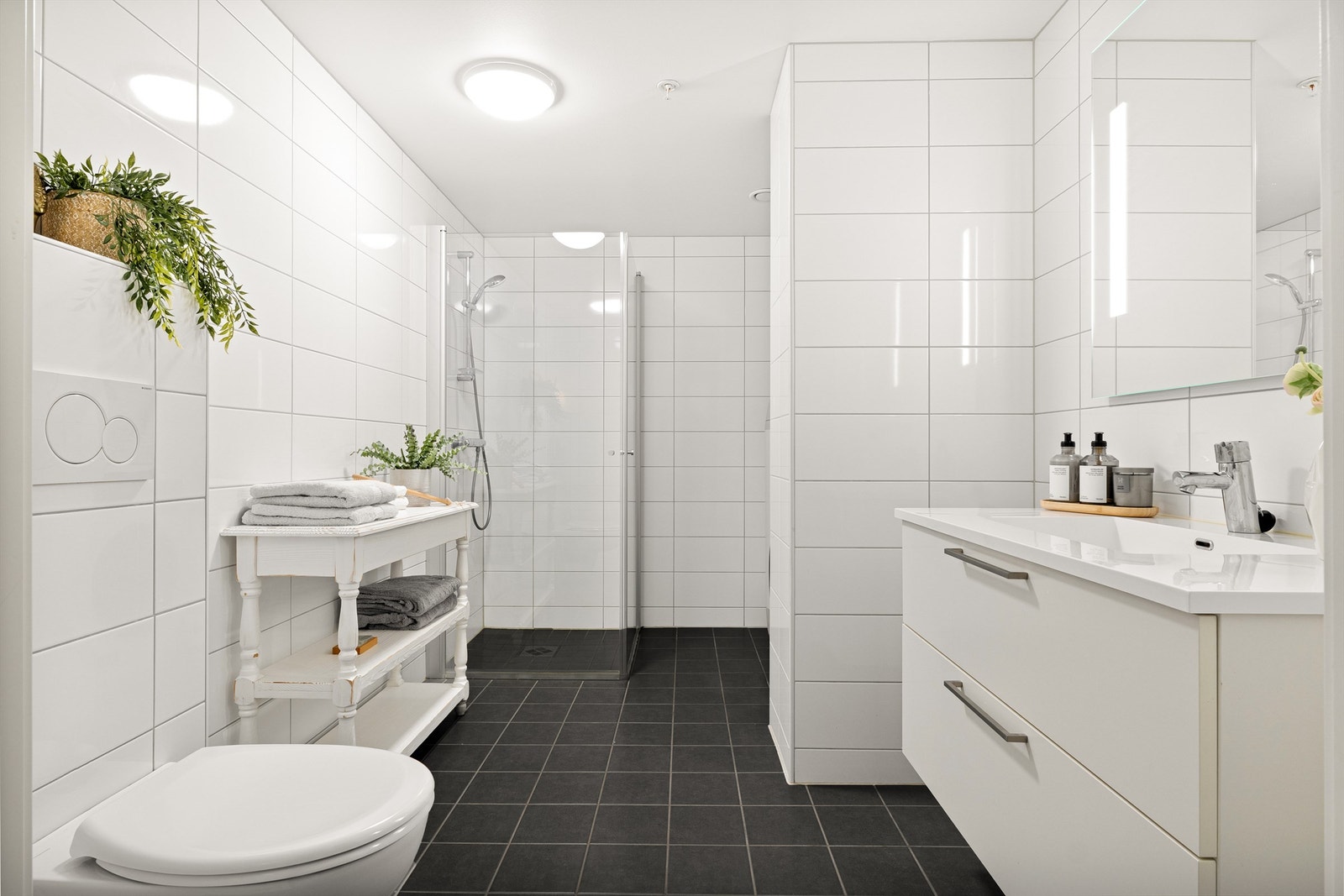 Tidløst og helfliset bad med varme i gulv. Badet er innredet med vegghengt toalett, dusj med glass foldedører, servant med møblement, speil og belysning. Galleribilde