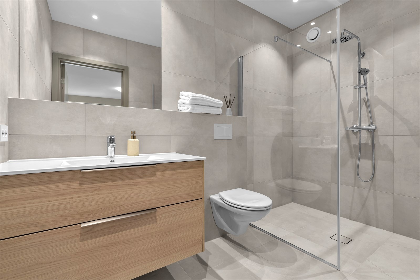 Baderommet inneholder vegghengt toalett, servant med underskuffer, speil og dusjhjørne med en vegg Galleribilde