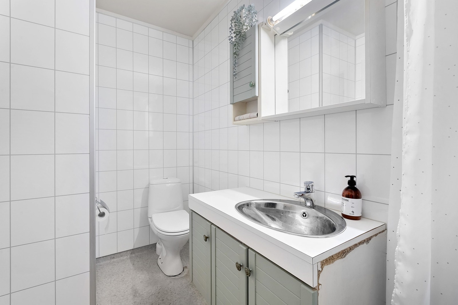 Badet er utstyrt med gulvmontert toalett, baderomsinnredning og dusj med forheng. Nyere vegger Galleribilde