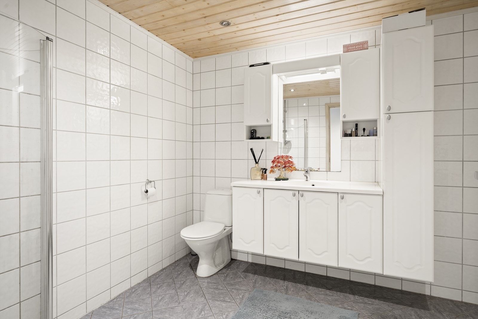 Romslig bad med flislagte overflater. Badet er utstyrt med toalett, skyllekum i stål, benkeskap med servant og foldbare dusjdører. Galleribilde