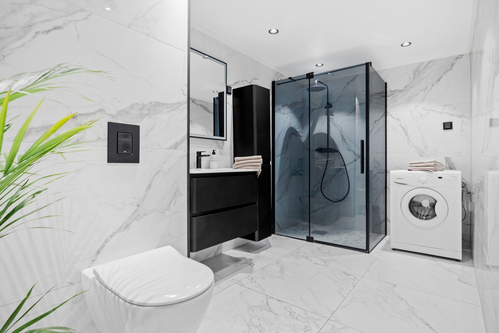 Badet er flislagt på vegger og gulv med varmekabler, dusjhjørne, vegghengt toalett og opplegg for vaskemaskin. Galleribilde