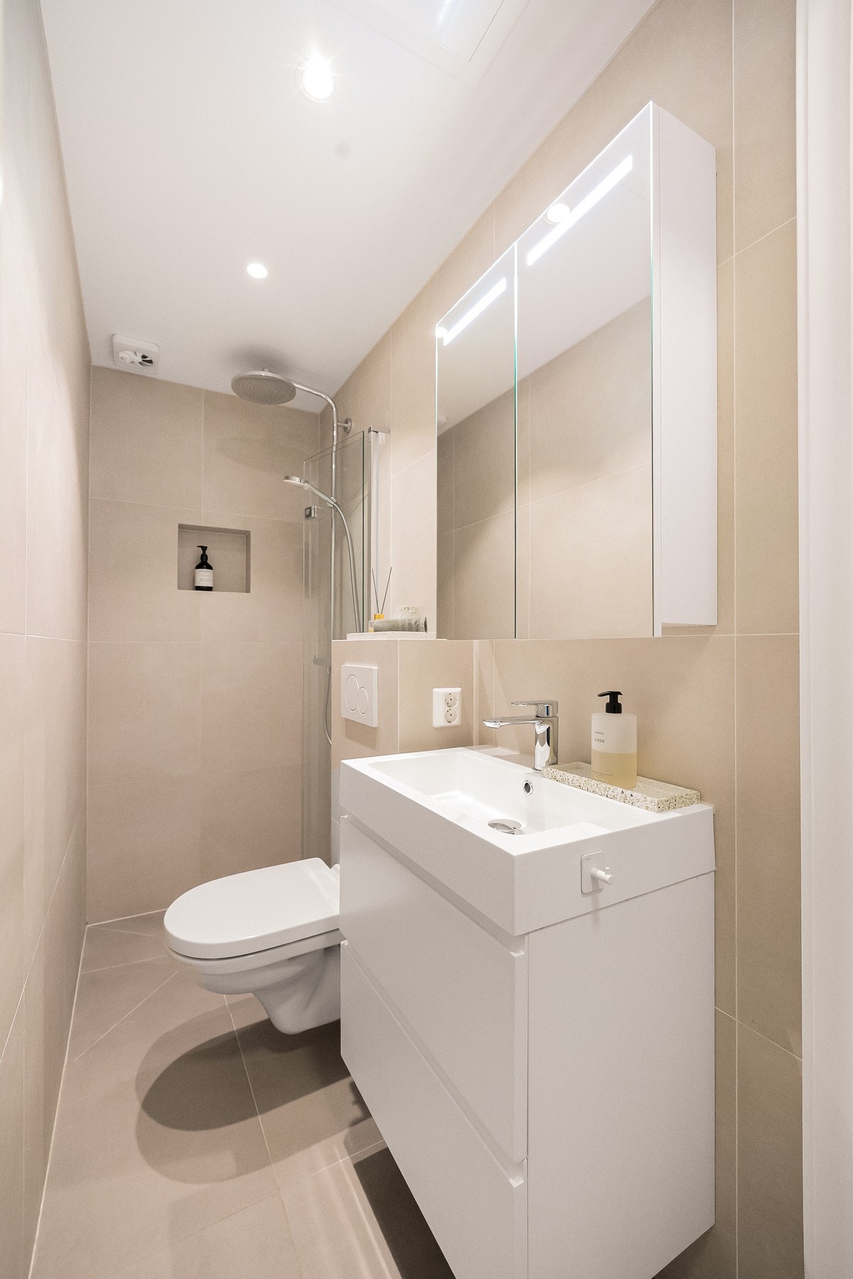 Videre har badet veggmontert servantskap med heldekkende vask og ett-greps armatur i krom, vegghengt speilskap med belysning samt innebygget sisterne med vegghengt toalett. Galleribilde