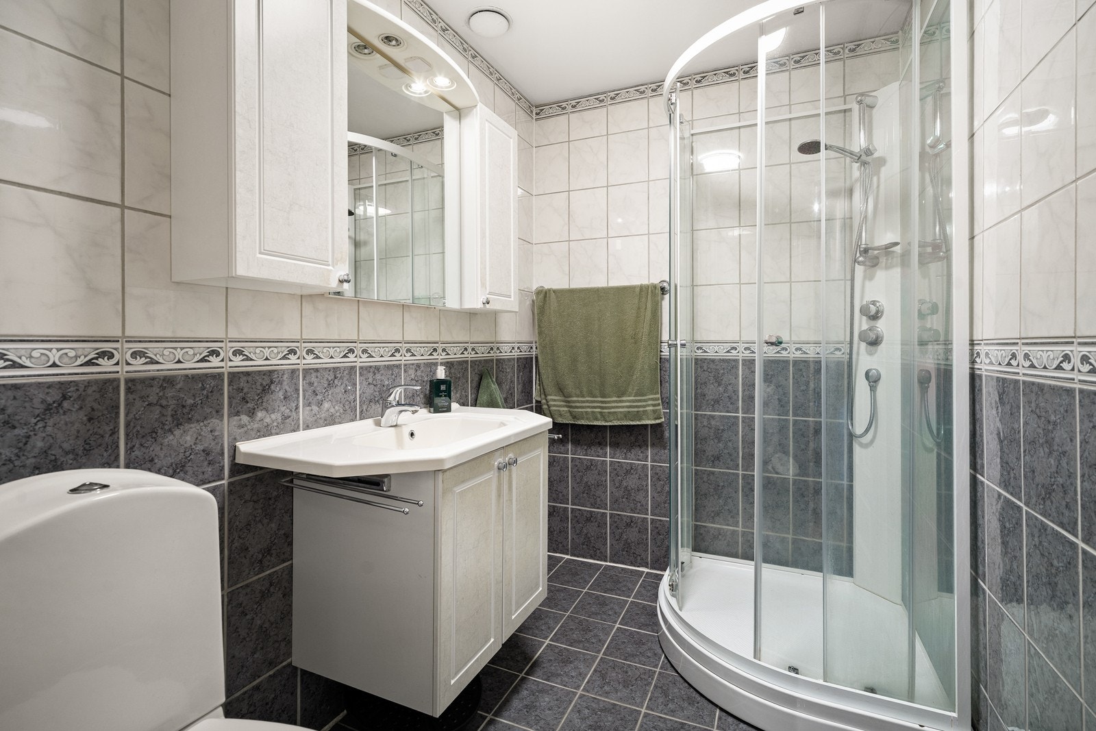 Bad i 2. etasje med flislagte overflater på gulv og vegger. Badet er innredet med baderomsmøbel med heldekkende servant med
profilerte, lyse fronter, servant armatur og overhengende speil med belysning. Galleribilde