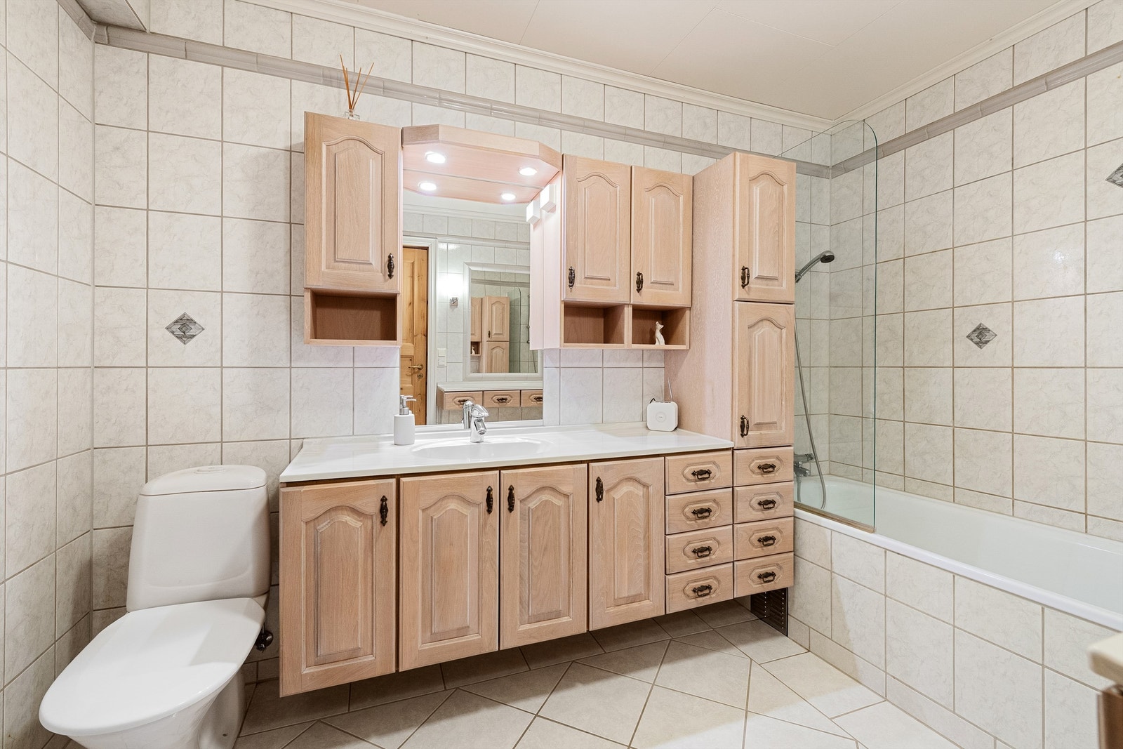 Stor baderomsinnredning med profilerte fronter og ovenpåliggende servant med ett-greps armatur. Speil med overlys og skap over servant. Innebygd badekar med dusjvegg og gulvstående toalett. Galleribilde