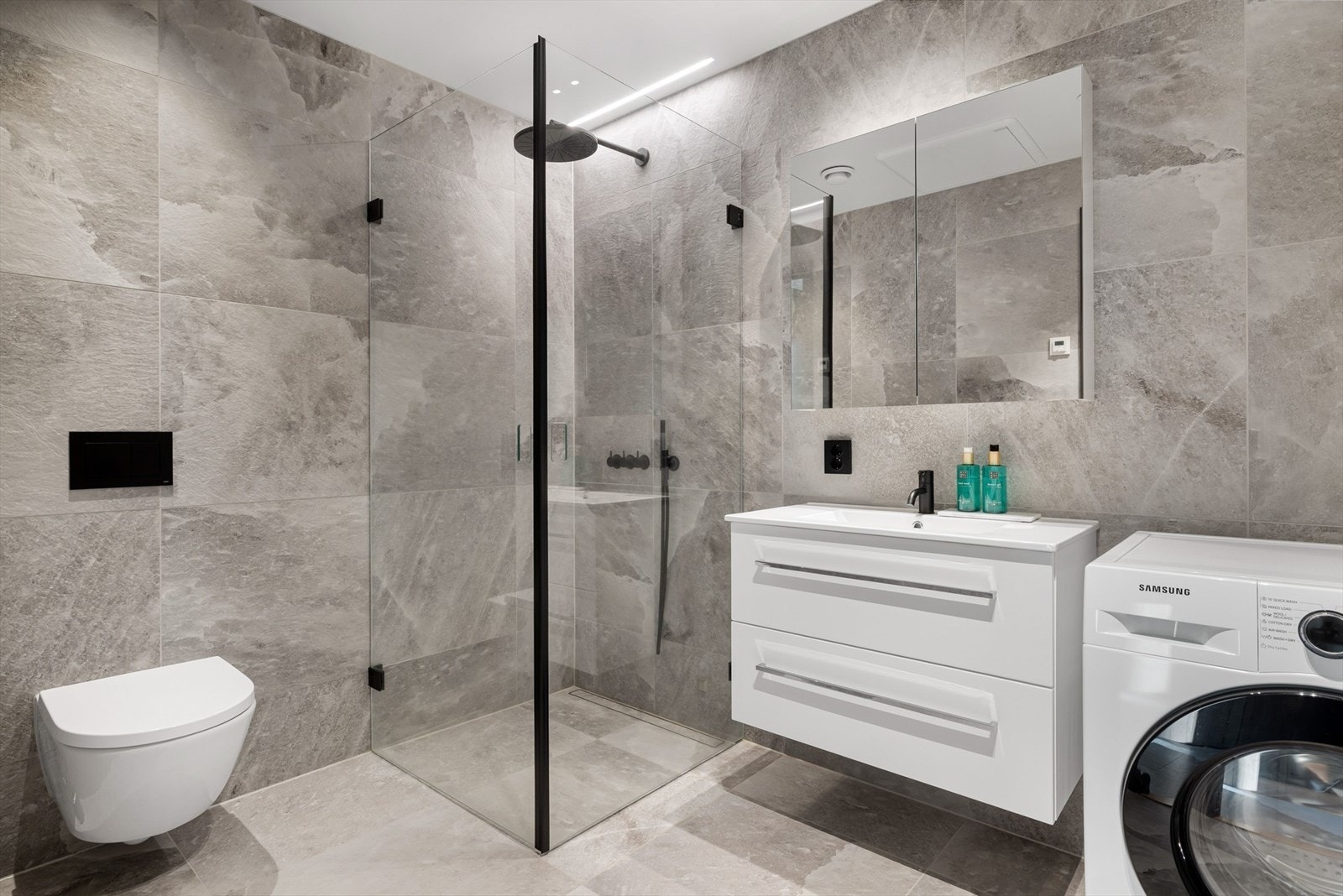 Lekkert baderom innredet med servant på innredning, vegghengt toalett, dusjhjørne med glassvegger og opplegg for vaskemaskin. Galleribilde