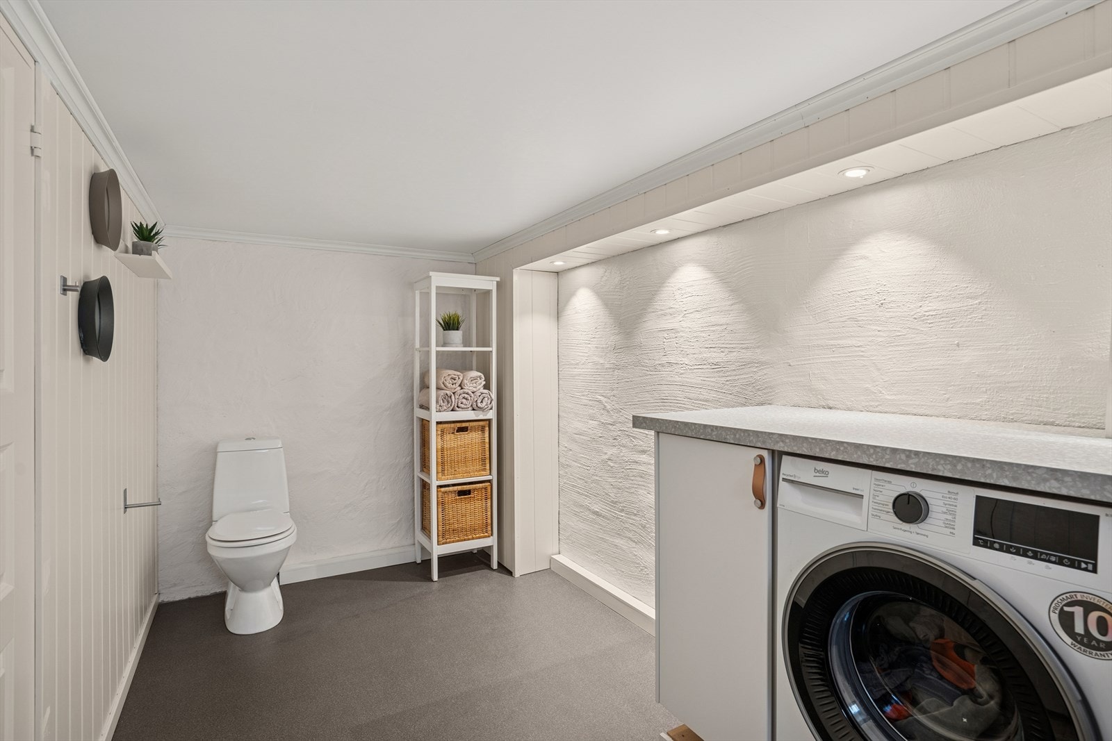 Vaskerom utstyrt med veggmontert panelovn, gulvmontert toalett, skyllekum i rustfritt stål og innredning med glatt front og benkeplate i høytrykkslaminat. Galleribilde