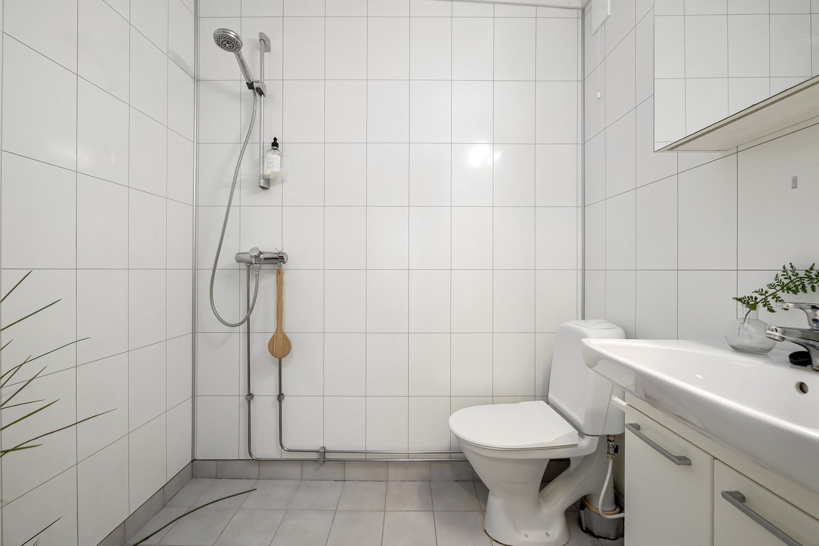 Flislagt baderom innredet med gulvstående toalett, vegghengt servant med skap under, speilskap med belysninger over og dusjsett Galleribilde