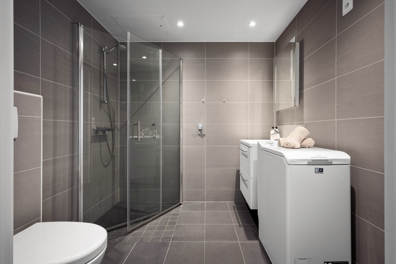 Moderne bad med fliser på gulv og vegger, dusjhjørne med glassdører, servant med innredning og vegghengt toalett. Galleribilde