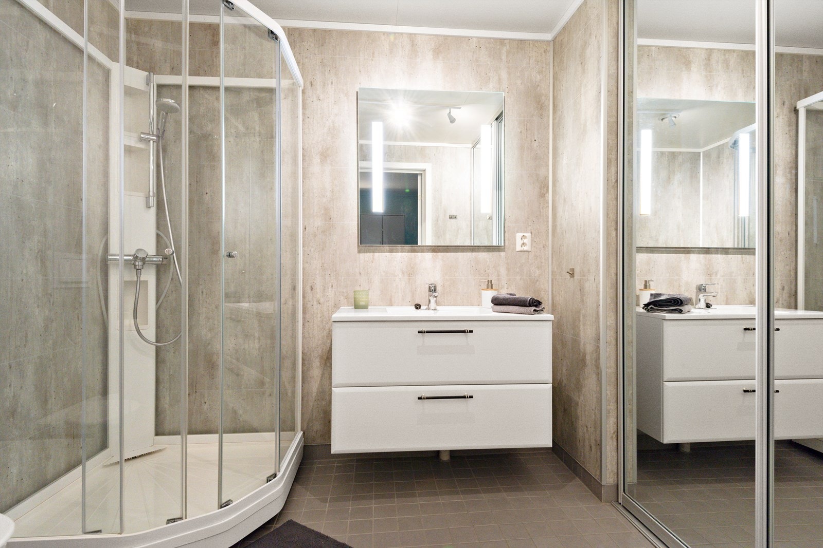 Badet med våtromsplater på vegg, flislagte gulv med varme, innredning, dusjkabinett, vegghengt toalett og skyvedører med speilfronter som skjermer mot vaskemaskin. Galleribilde