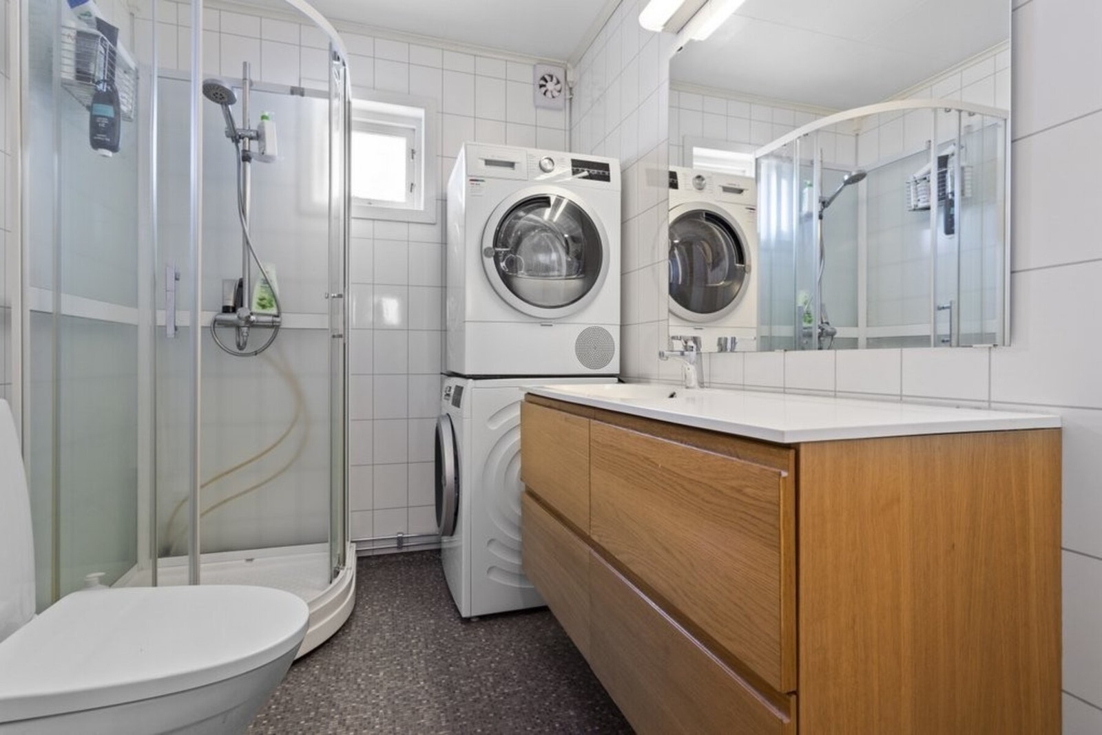 Pent bad med dusjkabinett, toalett og baderomsinnredning. Opplegg for både vaskemaskin og tørketrommel gir en praktisk og funksjonell løsning. Galleribilde