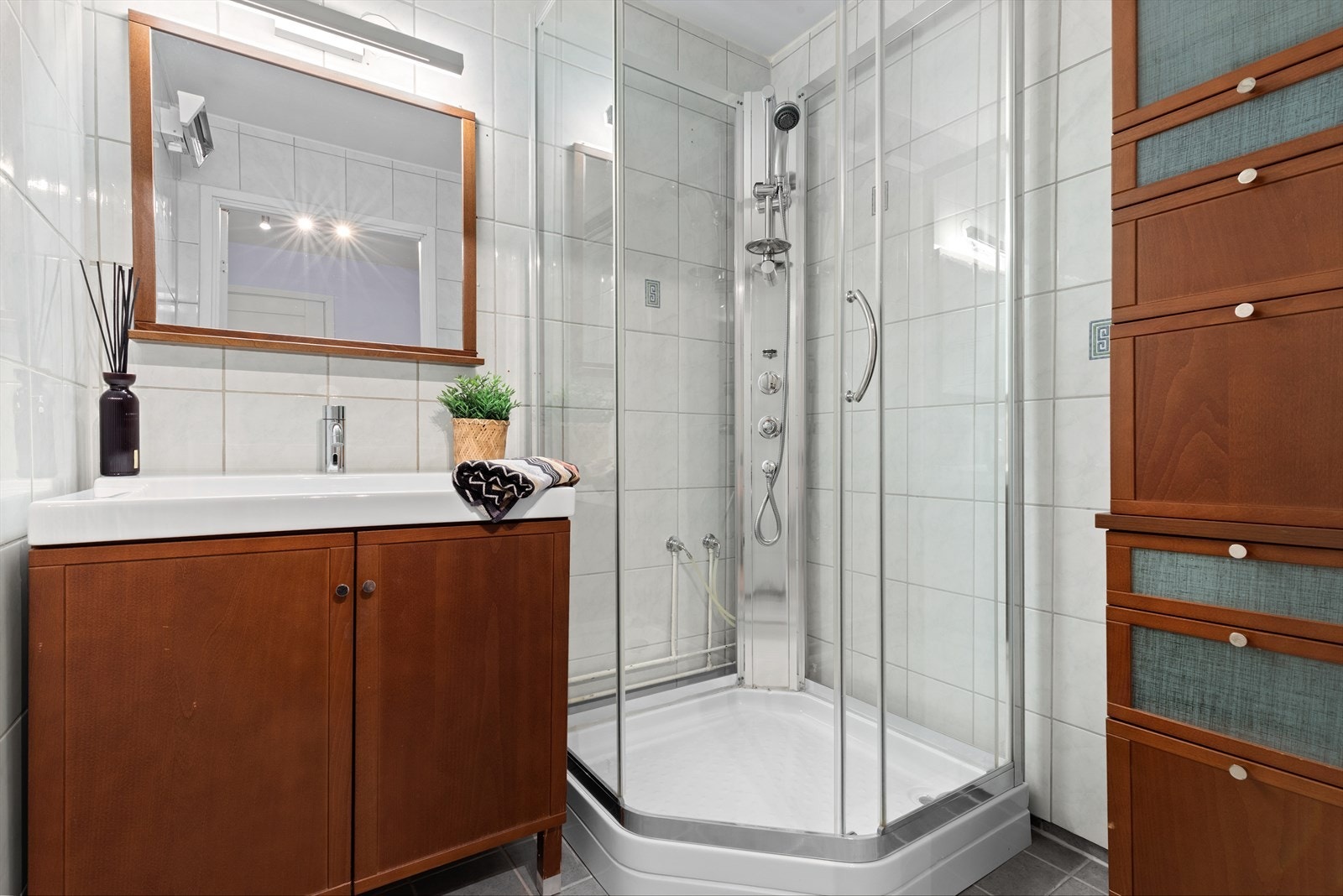 Badet har fliser på vegger og gulv, samt malt tak, og fremstår som lyst. Rommet er innredet med servant og dusjkabinett, som gir en praktisk og funksjonell dusjløsning. Galleribilde