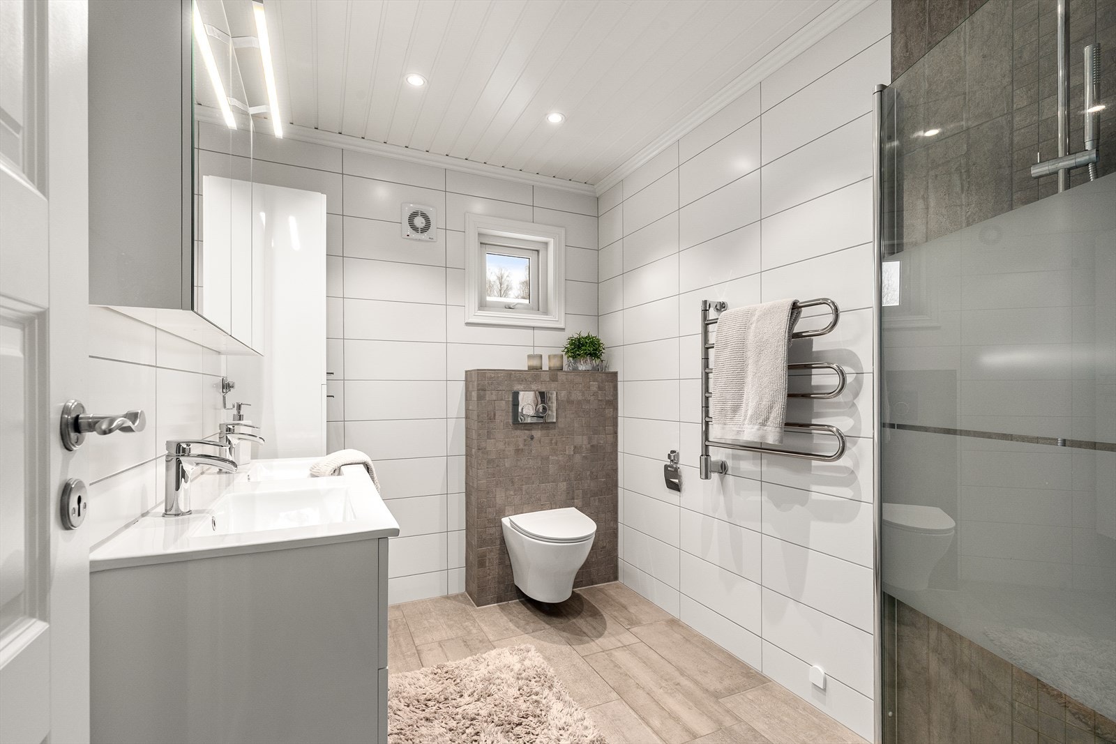 Delikat og moderne bad med flislagte overflater, vegghengt toalett, dusjnisje med glassvegg og praktisk baderomsinnredning med god oppbevaringsplass. Galleribilde