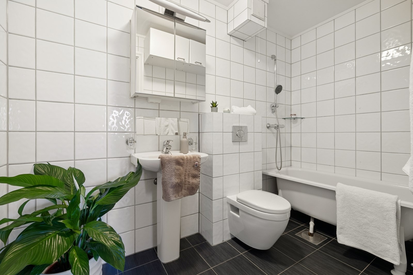 Flislagt baderom med varmekabler, badekar, vegghengt WC med innbygget sisterne, servant og speilskap med belysning. Galleribilde