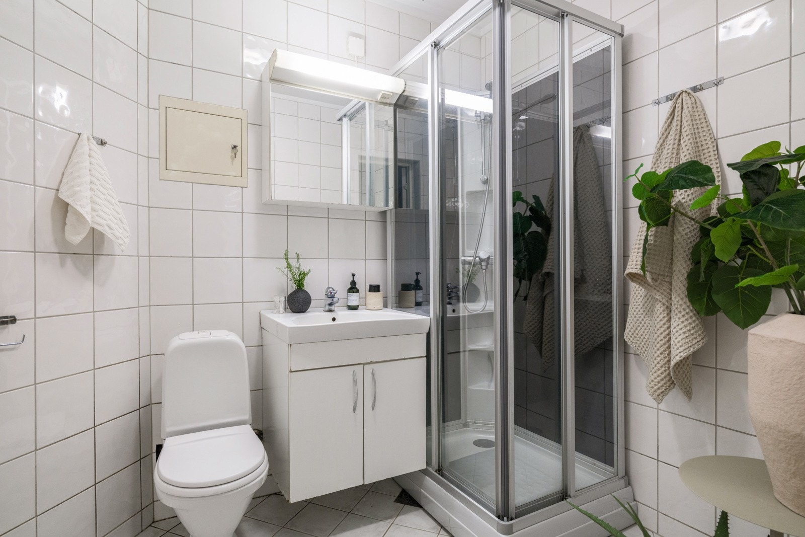 Baderommet har lyse fliser på gulv og vegger, varmekabler i gulv og eldre elektrisk avtrekksvifte. Rommet er utstyrt med servantskap, speilskap med belysning over, dusjkabinett og gulvstående gulv. Galleribilde