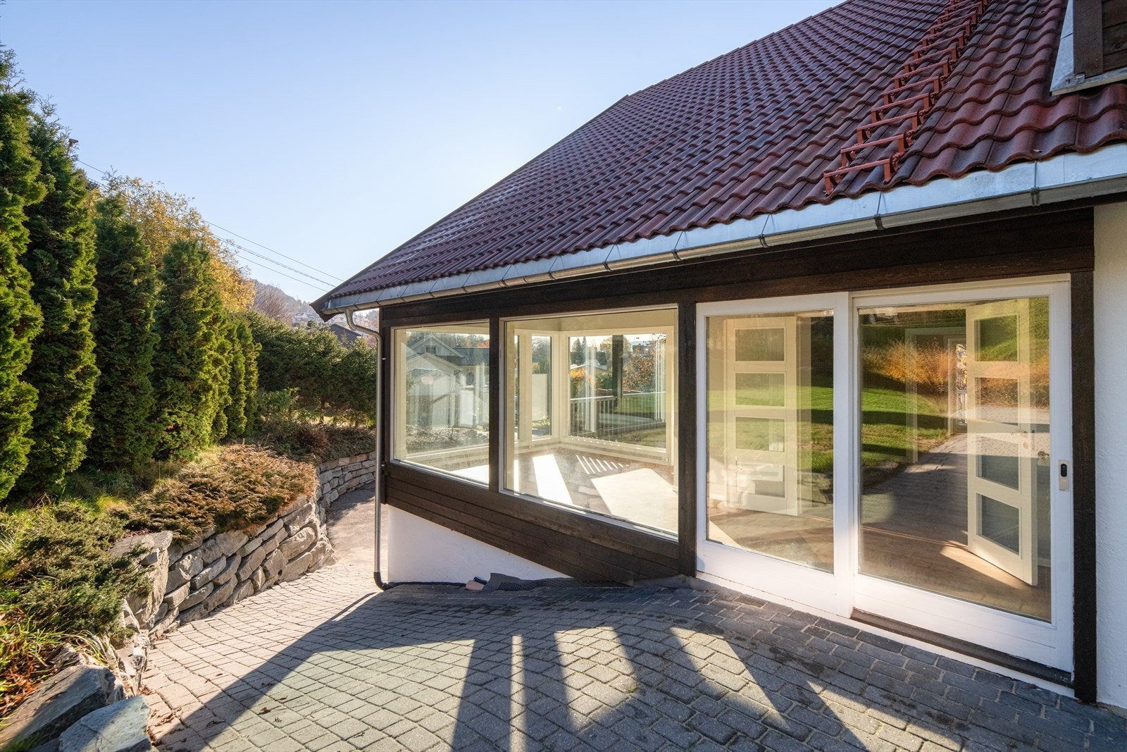 Tomta ligger i slakt skrånende terreng og er pent opparbeidet med asfaltert gårdsplass, forstøtningsmurer, grøntarealer med trær og prydbusker, og gangsti i hagen opp til en deilig uteplass. Galleribilde