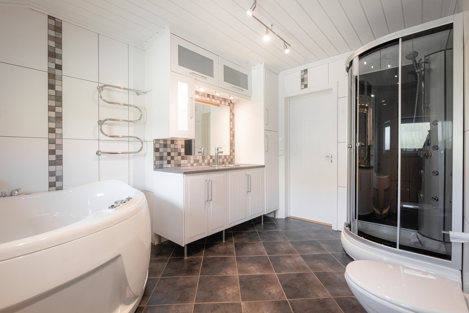 Her er innredning med meget god oppbevaringsplass, benkeplate med nedfelt servant og speil med overlys. Badet er også innredet med dusjkabinett, hjørnebadekar og veggmontert toalett. Galleribilde