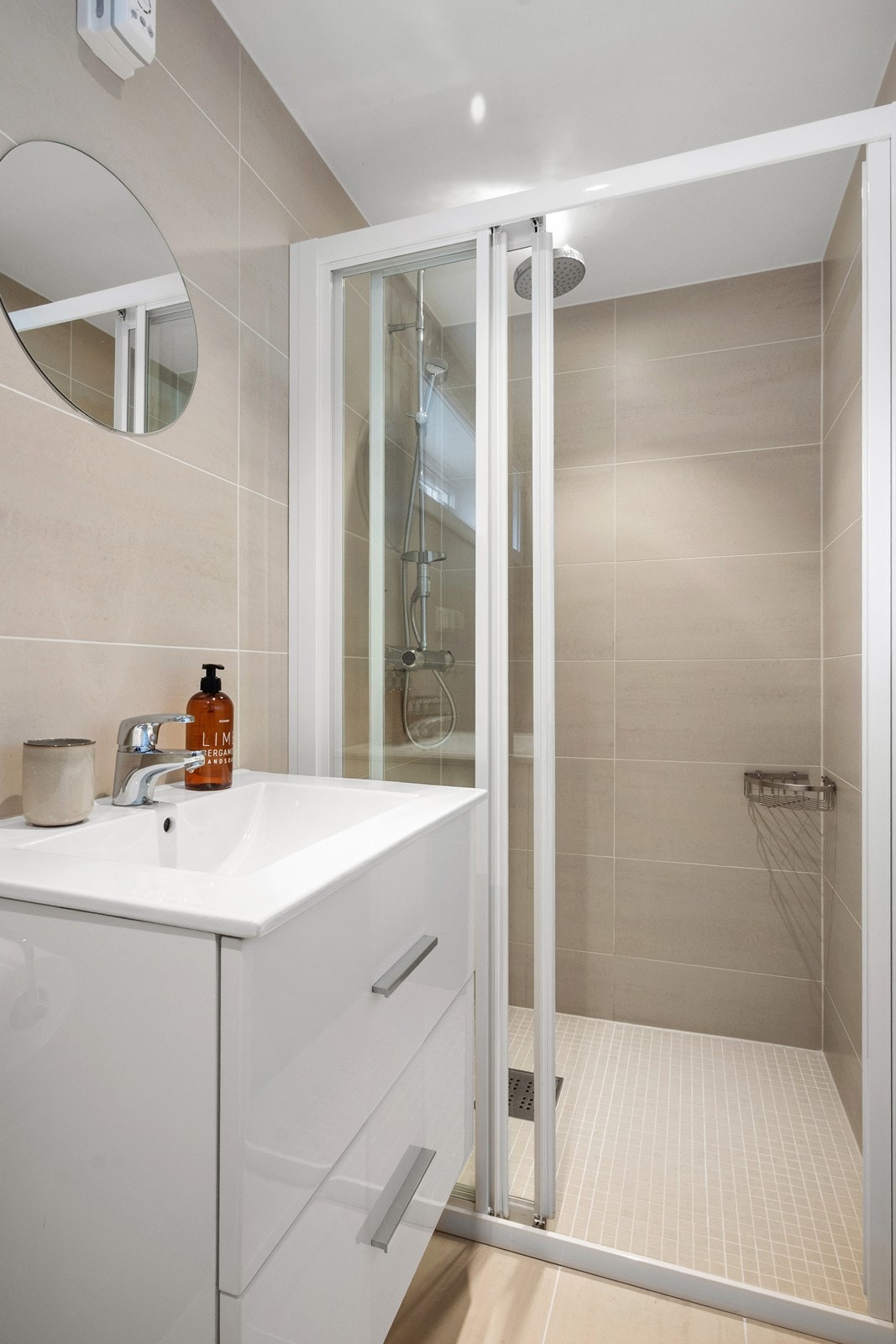 Rommet er flislagt i lyse, nøytrale toner og innredet med servant med skuffer, vegghengt toalett og dusjhjørne med mosaikkfliser og dusjdører i glass Galleribilde