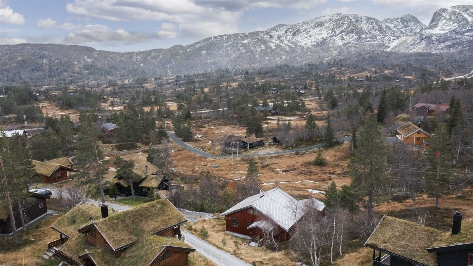 Luftfoto som viser hyttens plassering i et etablert hytteområde med storslått fjellandskap i bakgrunnen. Galleribilde
