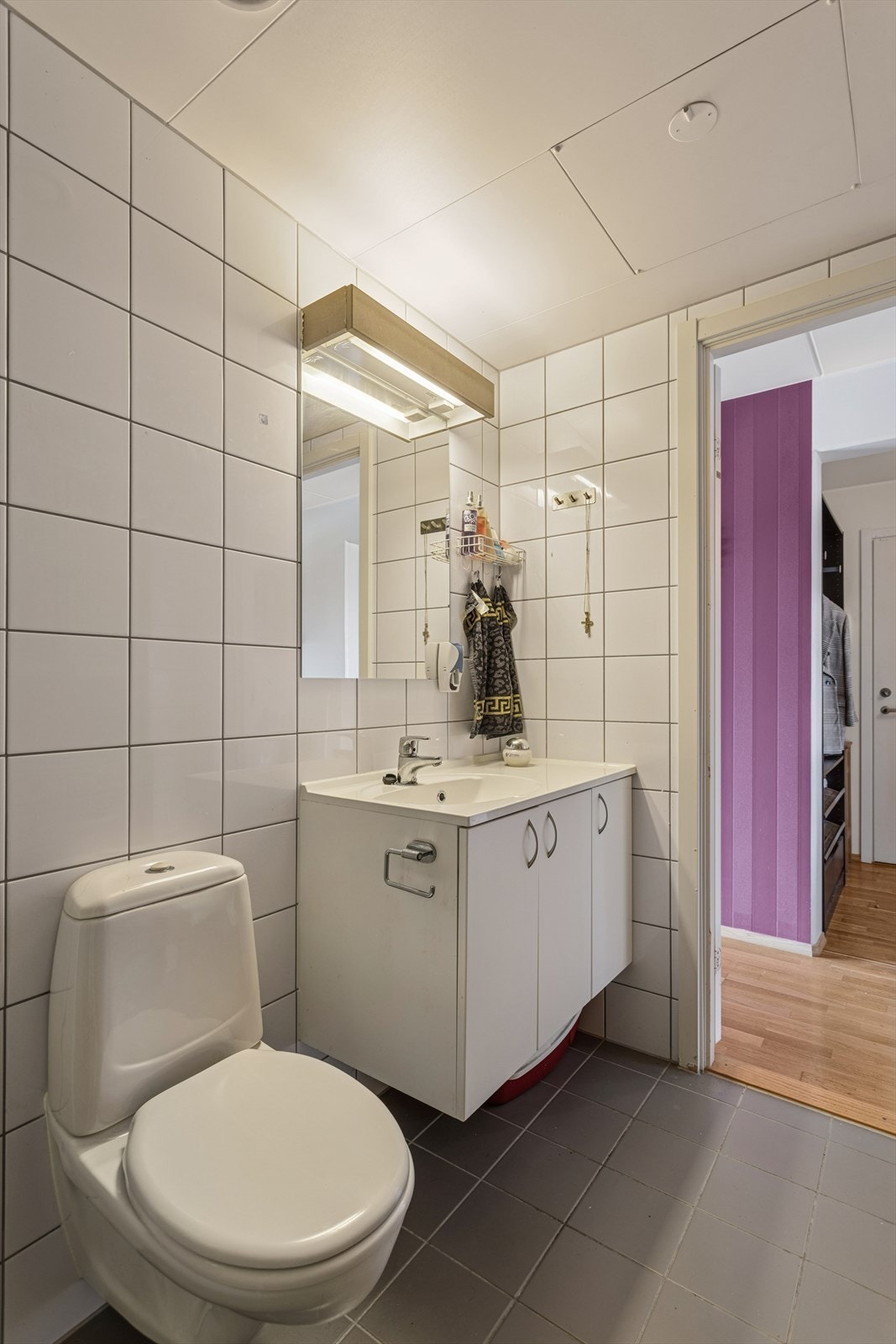 Badet er utstyrt med gulvmontert toalett, dusjhjørne med inn- og utadslående dører i herdet glass, samt servantseksjon med skapdører under. Galleribilde