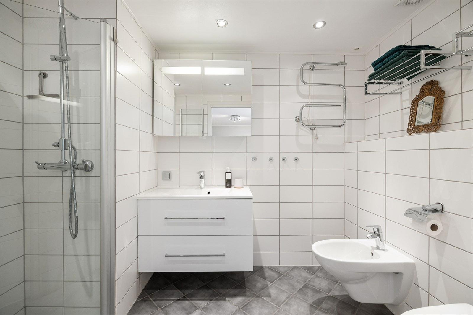 Badet er utstyrt med veggmontert toalett, bidé, dusjnisje med svingbare glassdører og servant med underskap. Galleribilde