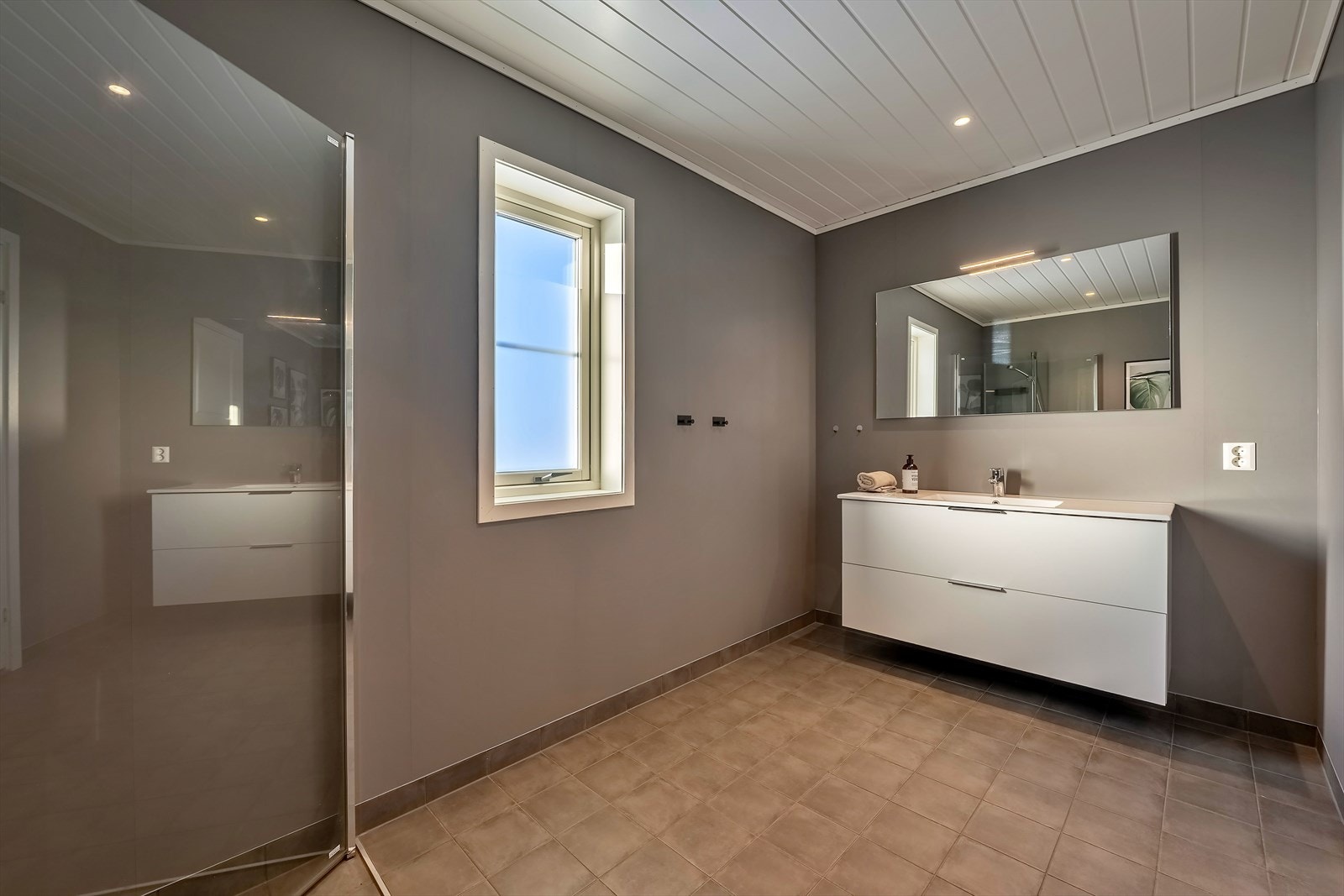 Romslig bad med flislagt gulv samt innredning med heldekkende servant, skuffer, speil og belysning Galleribilde