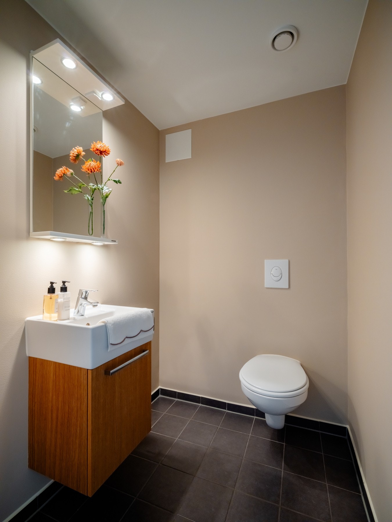 Stilrent toalettrom med flislagt gulv, vegghengt toalett og servant med speil og integrert belysning. Galleribilde