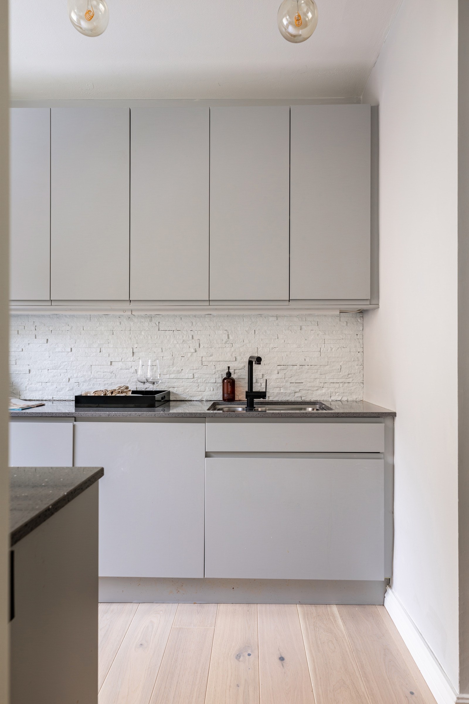 Løsningen består av glatte, lysegrå fronter med gripekant og benkeplate i grå stein med underlimt vaskekum og sort kran. Over benkeplaten er det montert belysning og lagt hvitmalte natursteinsfliser som backsplash. Galleribilde