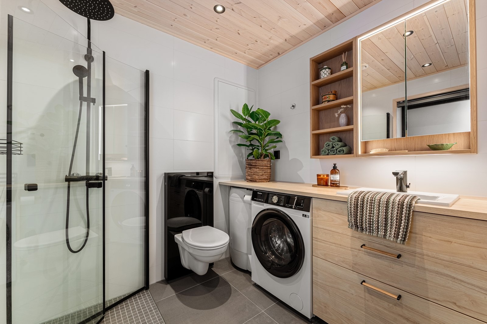 Badet er utstyrt med foldedører i dusjen, veggtoalett, opplegg for vaskemaskin og baderomsinnredning med servant, samt belyst speil Galleribilde