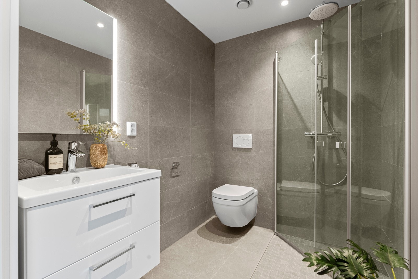 Badene ligger i hver sin etasje og har hjørnestilt dusjssone med utslagbare glassdører, vegghengt toalett og stilren servantinnredning med skuffer og speil med lys over. Det er lys grå 30x60 veggfliser og lys grå 60x60 gulvfliser. Galleribilde