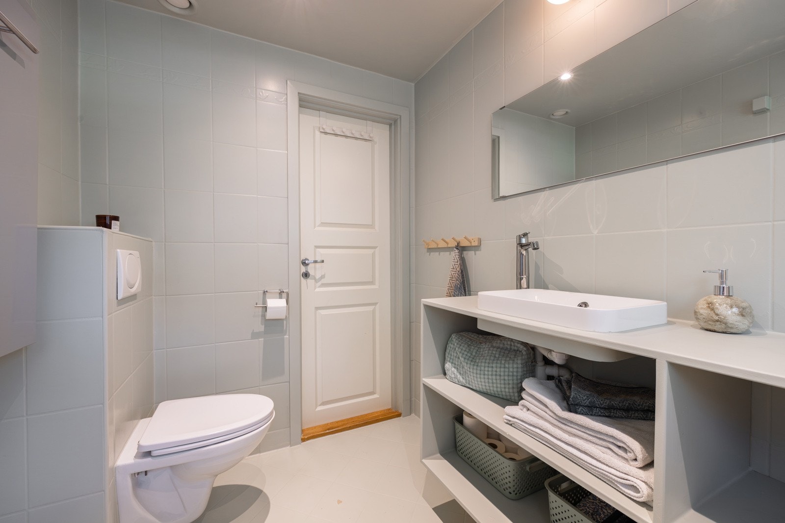 Plassbygget hylleløsning med ovenpåliggende servant og, dusjhjørne med innfellbar glassdør og veggspeil samt vegghengt toalett. God plass til vaskemaskin på badet, dersom man ønsker seg det, i tillegg har man tilgang til gratis fellesvaskeri i kjeller. Galleribilde