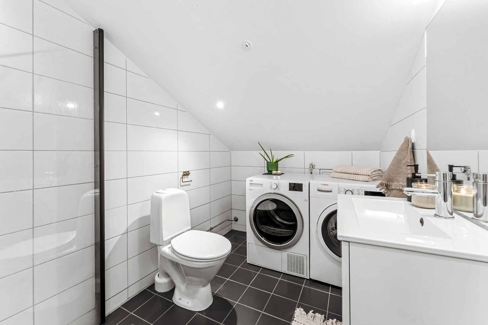 Videre er badet utstyrt med gulvmontert toalett, servantinnredning med skuffer og speil, samt dusjhjørne med innfellbare glassdører. Det er også opplegg for vaskemaskin og tørketrommel, med god plass til begge deler. Galleribilde