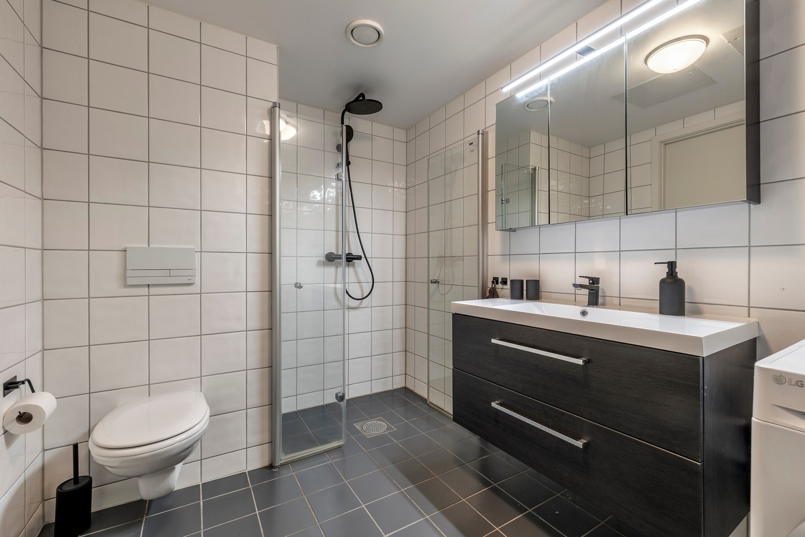 Innredet med servantinnredning, speil i dører på overskap på vegg over servant, vegghengt toalett og dusjhjørne med svingbare glassdører. Galleribilde