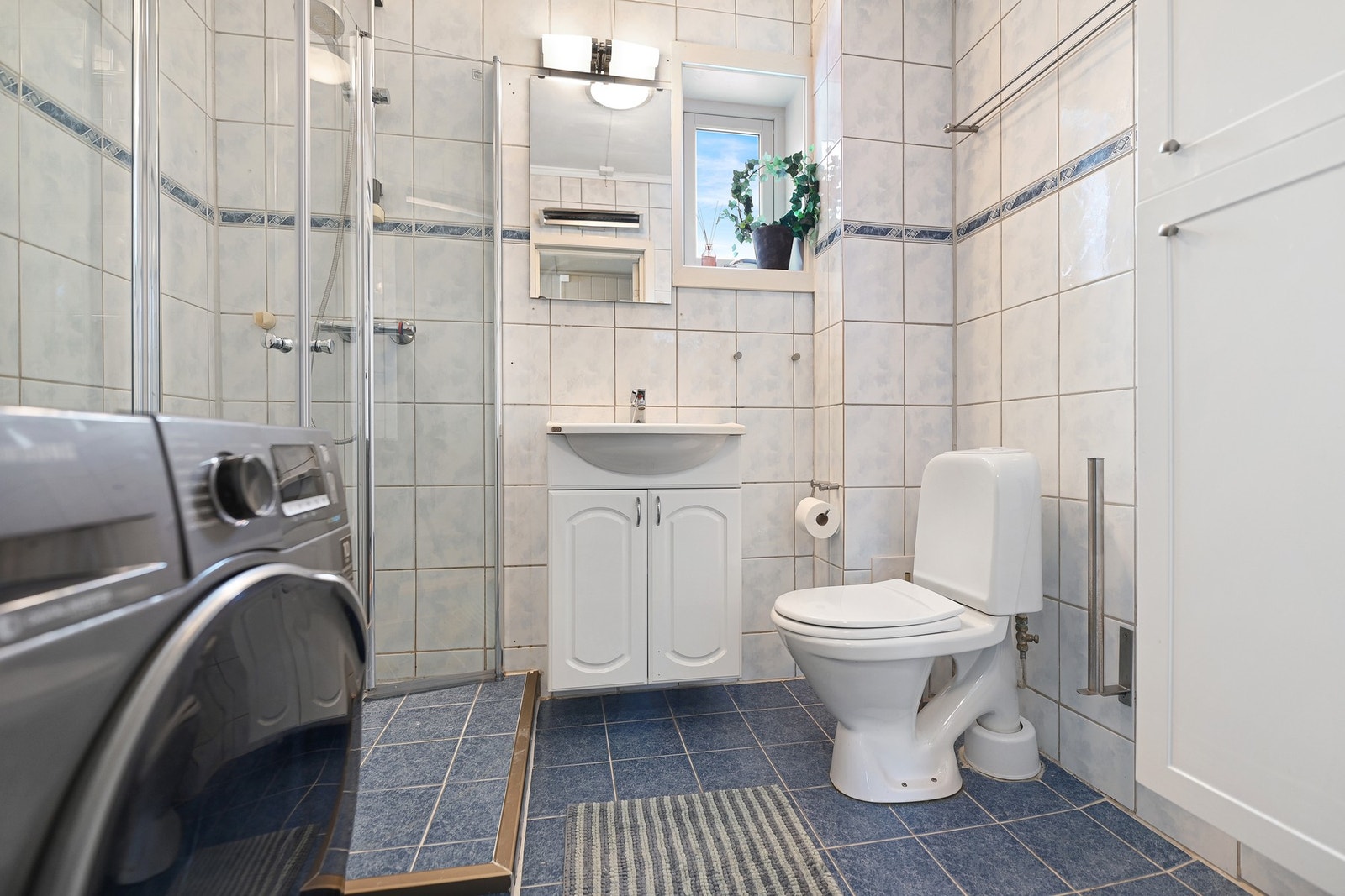 Badet ligger praktisk til fra gangen. Rommet er innredet med dusjhjørne, gulvstående wc, servantskap, speil, høyskap og opplegg for vaskemaskin. Det er naturlig ventilering. Galleribilde