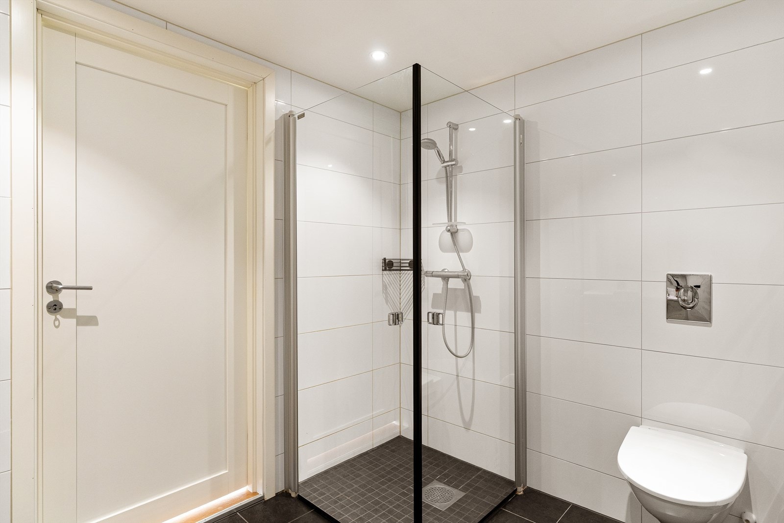 Badet har spottbelysning, dusjhjørne med glassvegger, vegghengt toalett, vaskeromsnisje og servant i badersomsinnredning. Galleribilde