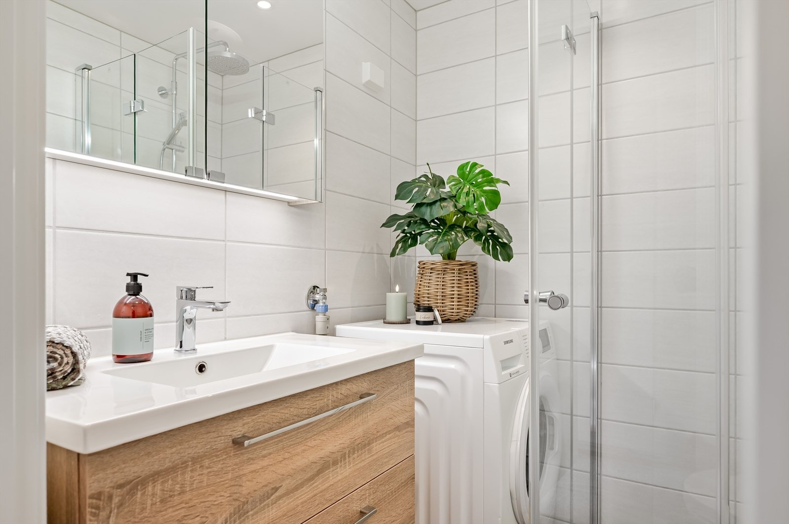 Moderne baderomsinnredning med skuffer og heldekkende servant, speilskap med belysning over. I hjørnet er det plass og opplegg for vaskemaskin. Galleribilde