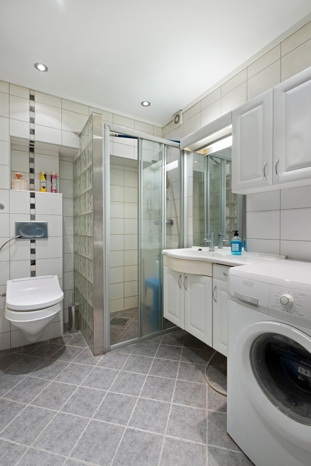 Lyst og pent bad med fliser på gulv og vegg, vegghengt toalett og plass til vaskemaskin. Galleribilde