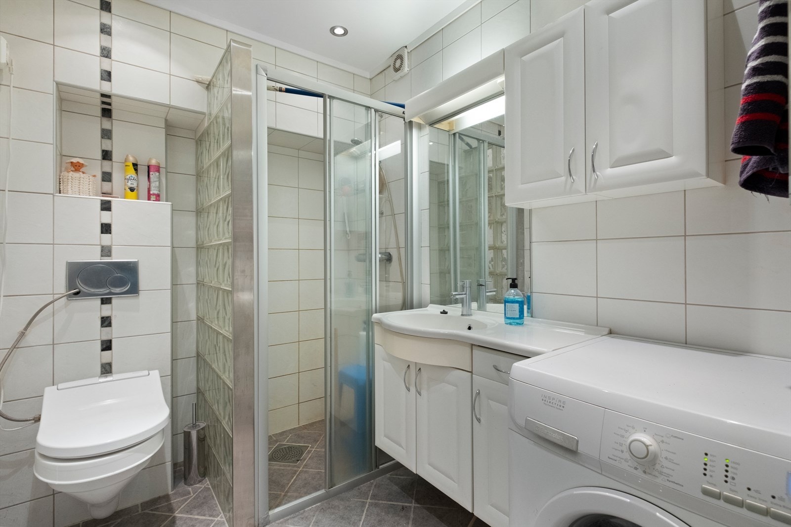 Lyst og pent bad med fliser på gulv og vegg, vegghengt toalett og plass til vaskemaskin. Galleribilde