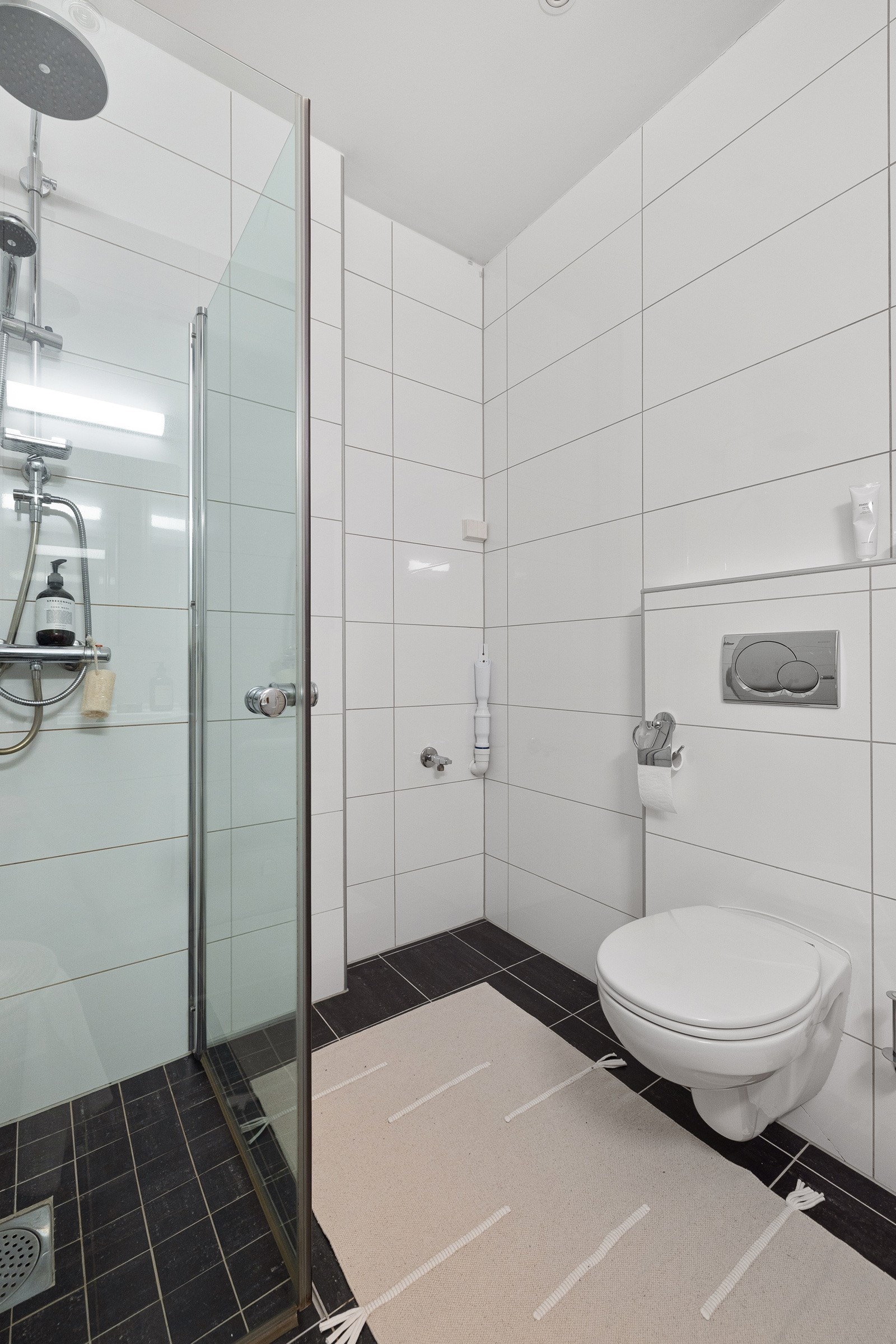 Badet er utstyr med gulvvarme, naturlig avtrekk, opplegg for vaskemaskin, rørfordelingsskap, stoppekran, veggmontert wc og dusjhjørne med glassdører og ettgreps dusjbatteri i rustfritt stål med to dusjhoder (håndholdt og regnfall). Galleribilde