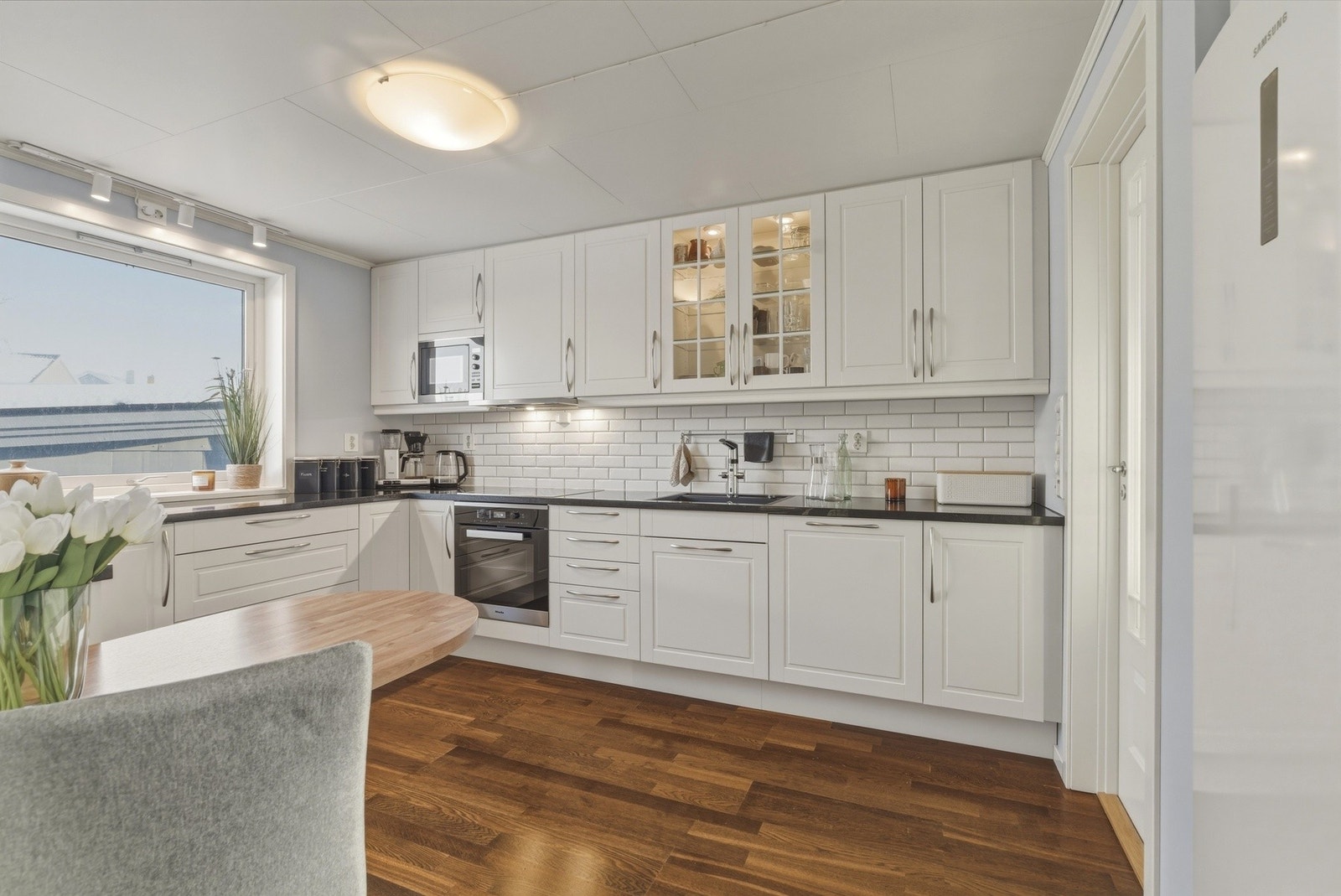 Flott og tidløst kjøkken fra Sigdal (2015) med hvite profilerte fronter og benkeplate i stein. Fliser på vegg over benk. Galleribilde