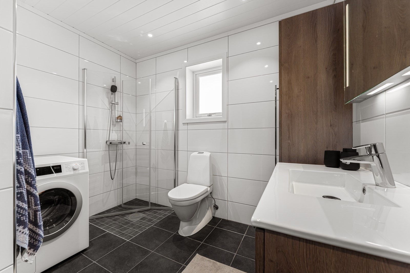 Flislagt bad med dusjhjørne, toalett, servant, speil med belysning og veggskap. Badet har også opplegg for vaskemaskin Galleribilde