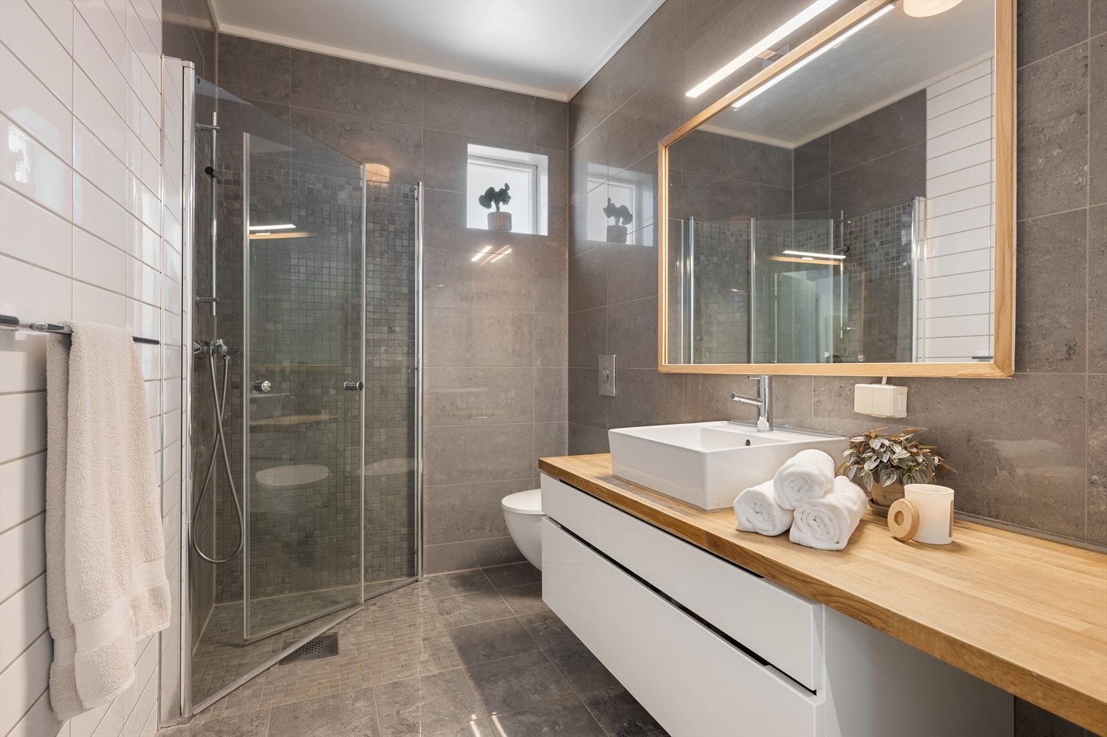 Meget lekkert bad av god størrelse. Pene mørke og lyse fliser på vegger og gulv. Badet er innredet med servant i innredning med 2 skuffer, samt benkeplate. Speil med belysning over. Vegghengt toalett. Dusj med innfellbare glassdører Galleribilde