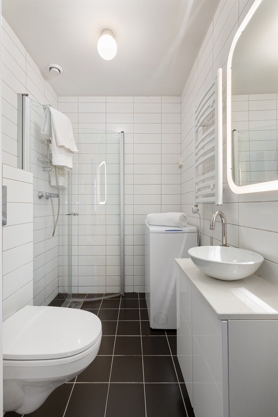 Pent flislagt bad med servant på steinplate med underskap, speil med lys, håndkletørker, veggmontert wc og veggmonterte dusjdører. Galleribilde