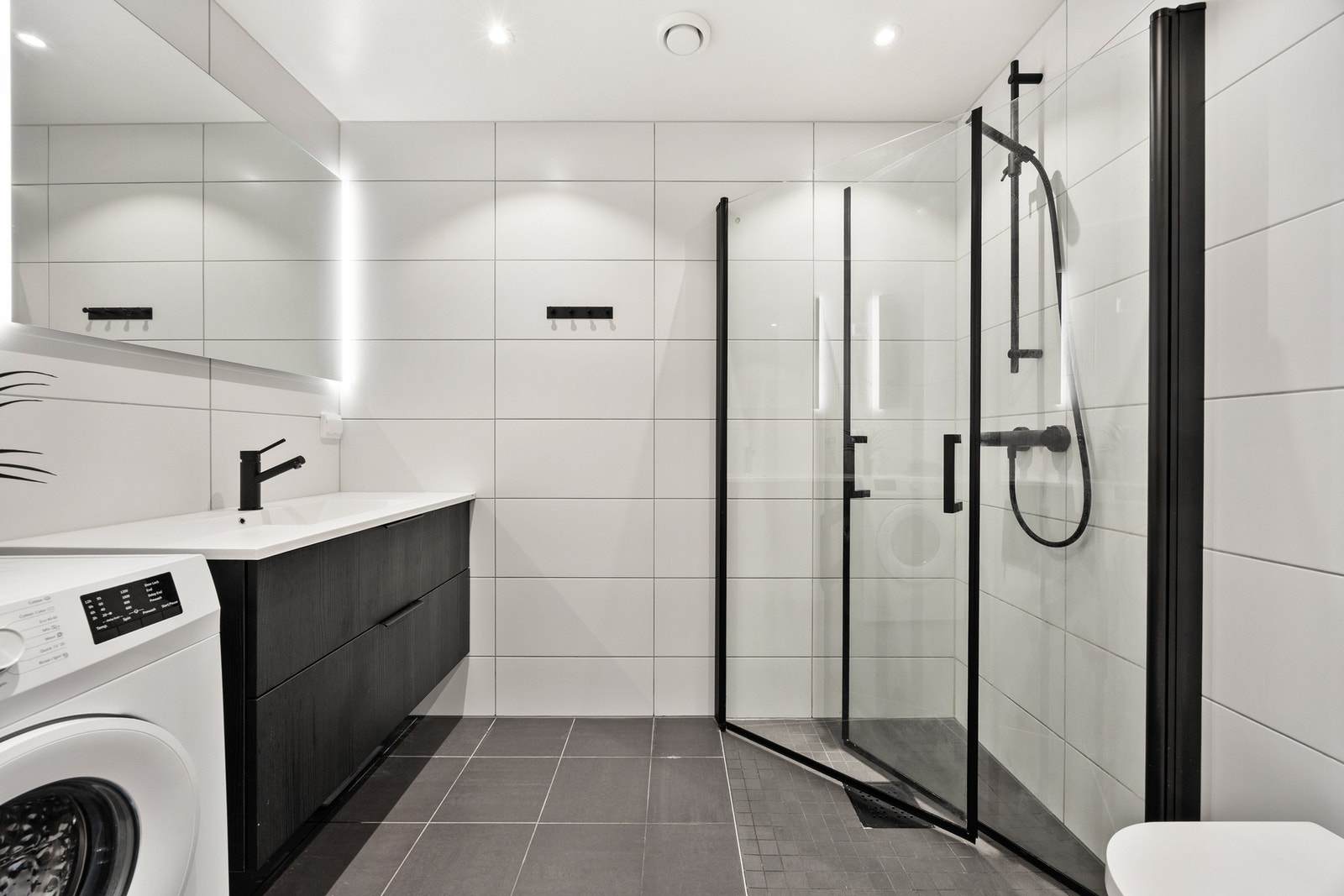 Lyst, pent og helfliset bad med gulvvarme. Badet har dusj med dører i herdet glass, servant, veggfestet klosett, speil med belysning over og opplegg for vaskemaskin. Her har du rikelig med rom for oppbevaring i skuffeseksjon under servant Galleribilde