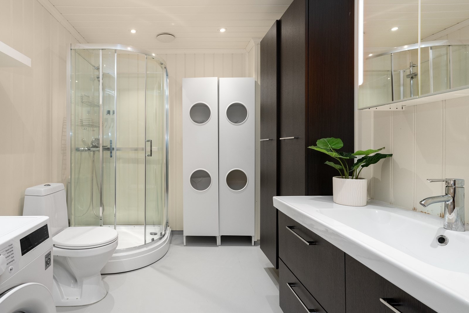 Leiligheten har et romslig bad som også fungerer som vaskerom, med opplegg for og plass til vaskemaskin. Innredningen i mørk utførelse har godt med oppbevaringsplass, samt at det også er speilskap. Badet er også innredet med dusjkabinett og toalett. Galleribilde