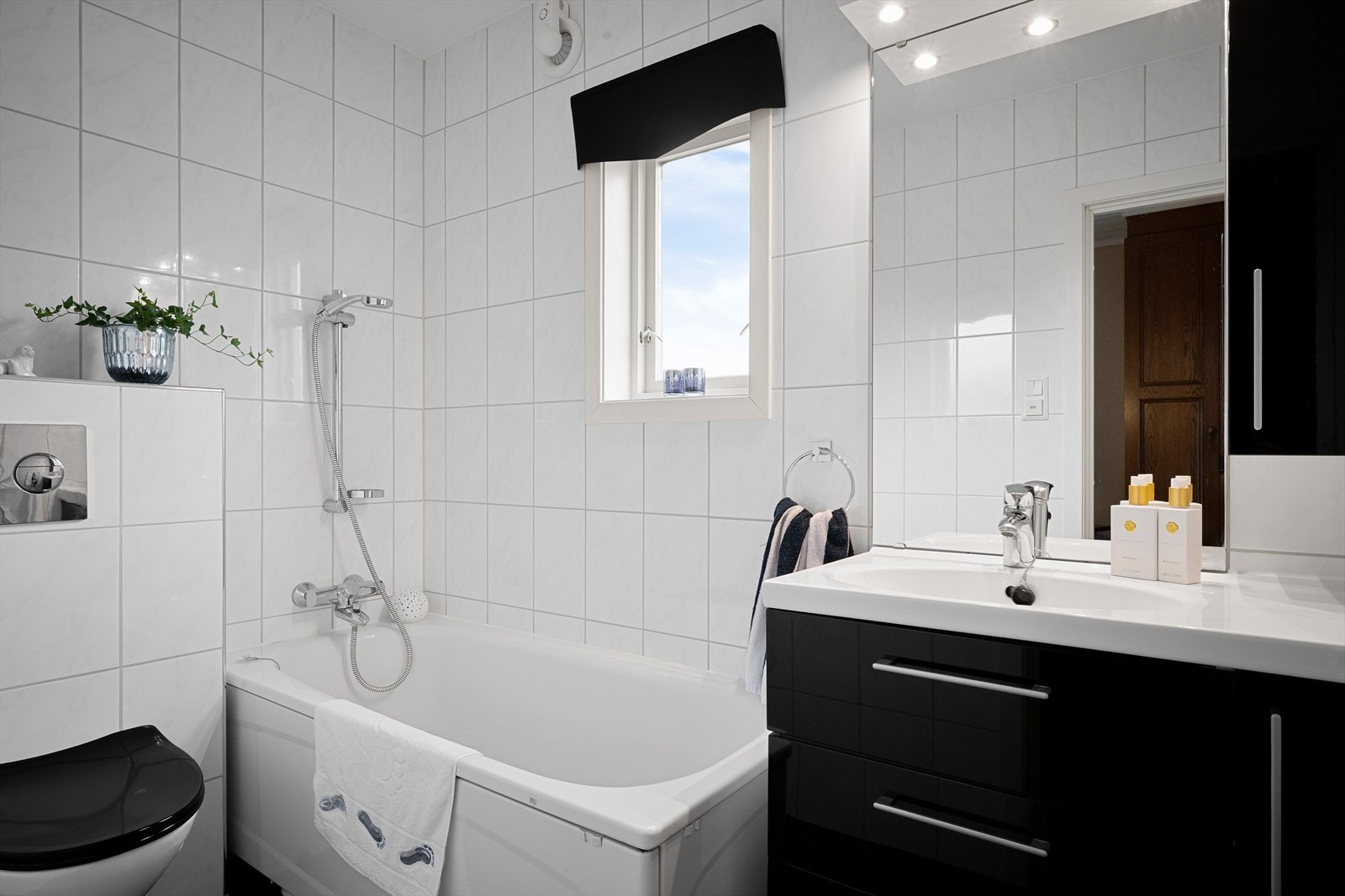 Helfliset bad med spotter i tak. Badekar, sort seksjon med vask/benk, overskap og speil på vegg. Veggmontert toalett. Galleribilde